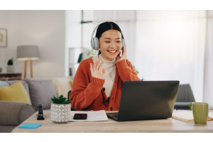 Imagem de uma jovem mulher sentada em uma mesa, interagindo com alguém através de uma videochamada em seu notebook. Ela está usando um fone de ouvido e sorrindo, enquanto acena com a mão direita. Ao fundo, é possível ver um ambiente de sala de estar. 