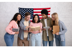 A imagem mostra um grupo de cinco jovens sorridentes, segurando livros e tablets, em frente a uma bandeira dos Estados Unidos. Eles parecem empolgados e interagindo em um ambiente descontraído, sugerindo uma atividade de aprendizado ou discussão em grupo.