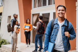 um jovem estudante com mochila e material de estudo sorri com confiança. Ao fundo, outros alunos interagem em um ambiente educacional. Os cursos profissionalizantes oferecem conhecimento prático e direcionado para o sucesso profissional. 