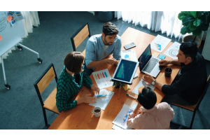 Quatro pessoas estão reunidas ao redor de uma mesa de madeira em um ambiente de escritório, discutindo gráficos e relatórios exibidos em um laptop e em papéis espalhados pela mesa. A cena sugere uma reunião colaborativa de análise de dados.