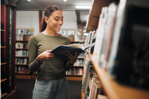 A imagem retrata uma jovem sorrindo enquanto lê um livro em uma biblioteca. Ela está de pé perto de uma estante, vestindo uma blusa verde e jeans. Ela lê para alavancar sua carreira.