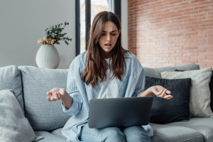  Na imagem, uma mulher, sentada em um sofá, expressa confusão ou frustração enquanto olha para a tela de seu laptop, gesticulando com as mãos abertas em sinal de dúvida.