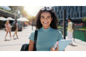 A imagem mostra uma jovem sorridente ao ar livre, carregando cadernos e uma mochila. Ela usa uma blusa verde clara e óculos de sol no cabelo, em um ambiente ensolarado e urbano.