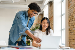 A imagem mostra dois jovens em um ambiente de escritório, onde um homem está orientando uma mulher que usa um laptop. Ambos estão sorrindo, sugerindo uma interação colaborativa e amigável.