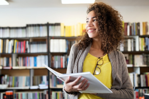 Jovem sorridente em uma biblioteca, segurando um livro aberto, transmitindo uma atmosfera de confiança e alegria.