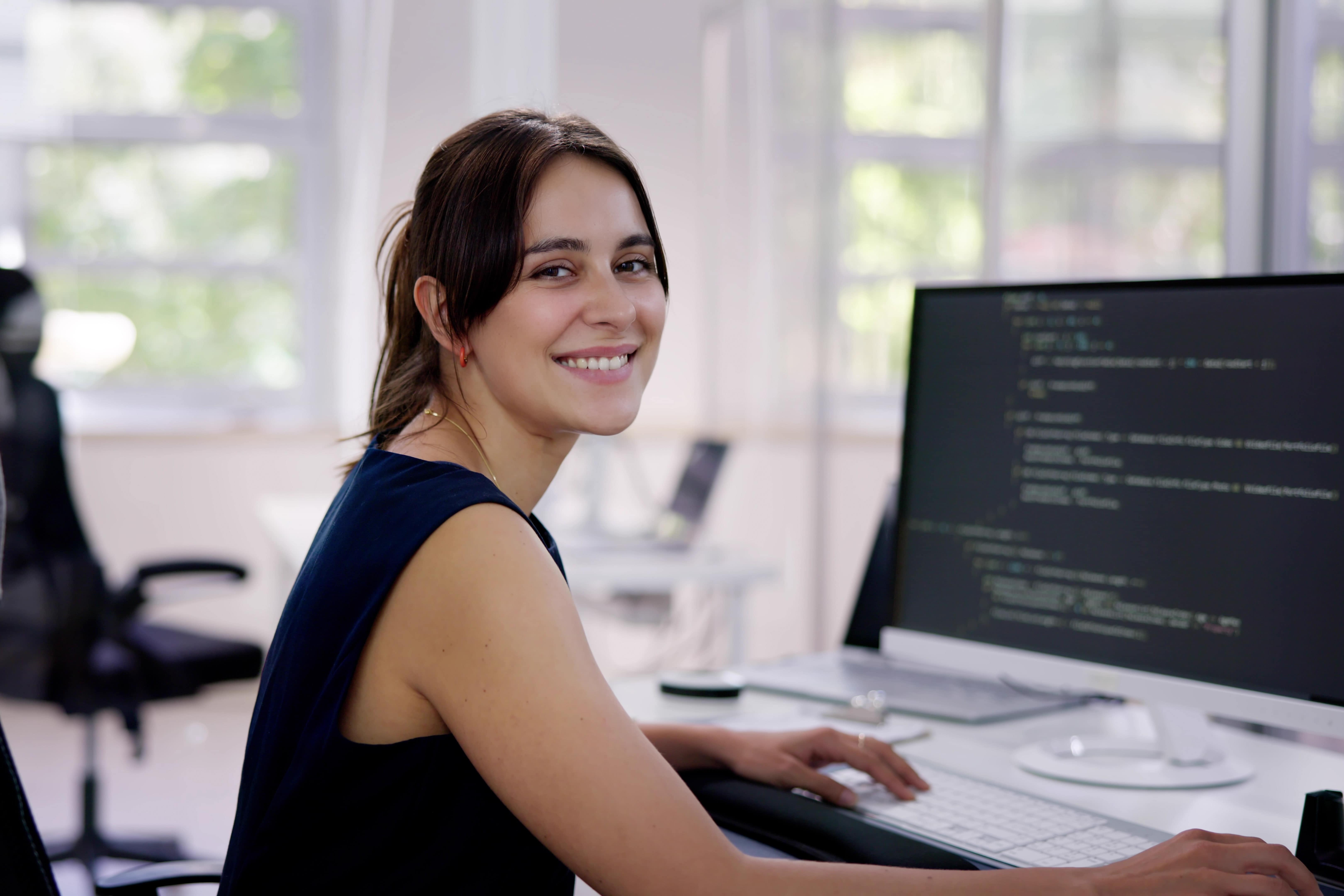  Imagem de uma mulher branca, sorrindo, trabalhando na área de análise e desenvolvimento de sistemas em um escritório.