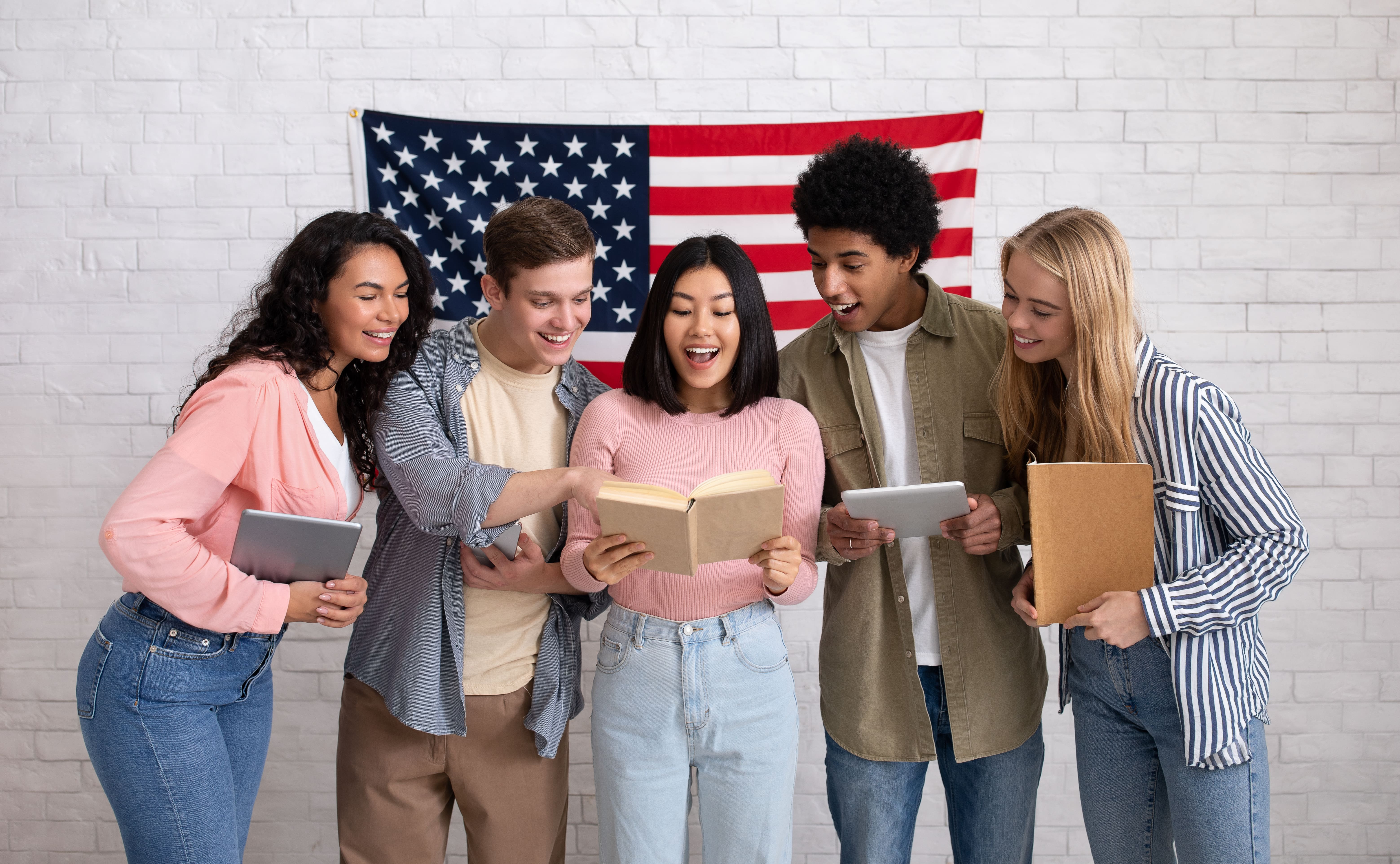 A imagem mostra um grupo de cinco jovens sorridentes, segurando livros e tablets, em frente a uma bandeira dos Estados Unidos. Eles parecem empolgados e interagindo em um ambiente descontraído, sugerindo uma atividade de aprendizado ou discussão em grupo.