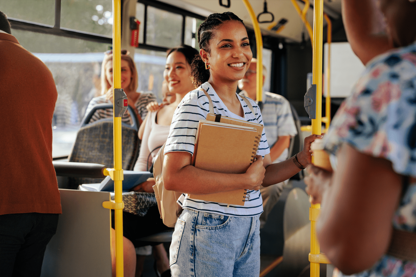 Uma jovem estudante sorridente, carregando cadernos e mochilas, está de pé em um ônibus, interagindo com outra pessoa enquanto se prepara para seu dia de trabalho. Ao fundo, outros passageiros estão sentados.