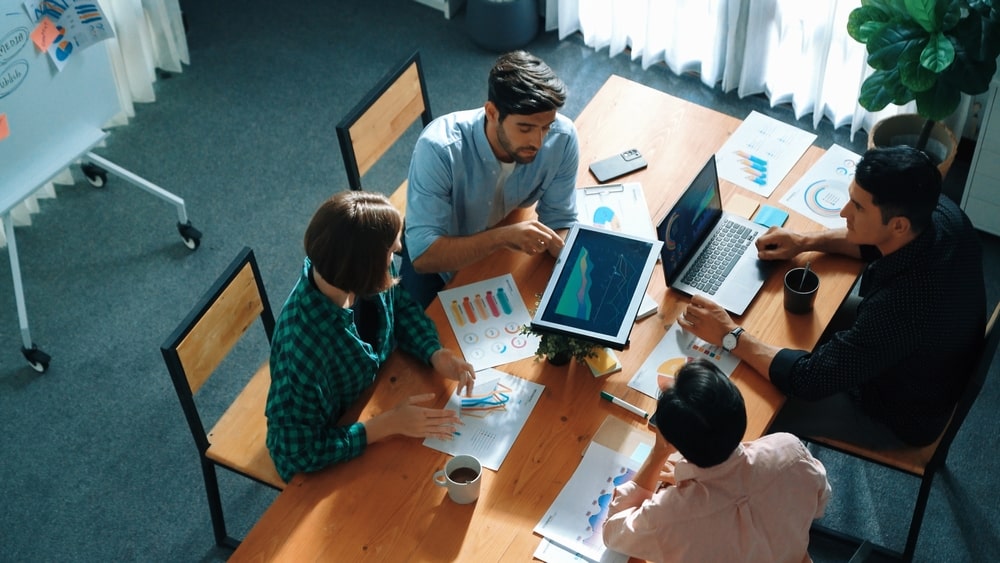 Quatro pessoas estão reunidas ao redor de uma mesa de madeira em um ambiente de escritório, discutindo gráficos e relatórios exibidos em um laptop e em papéis espalhados pela mesa. A cena sugere uma reunião colaborativa de análise de dados.