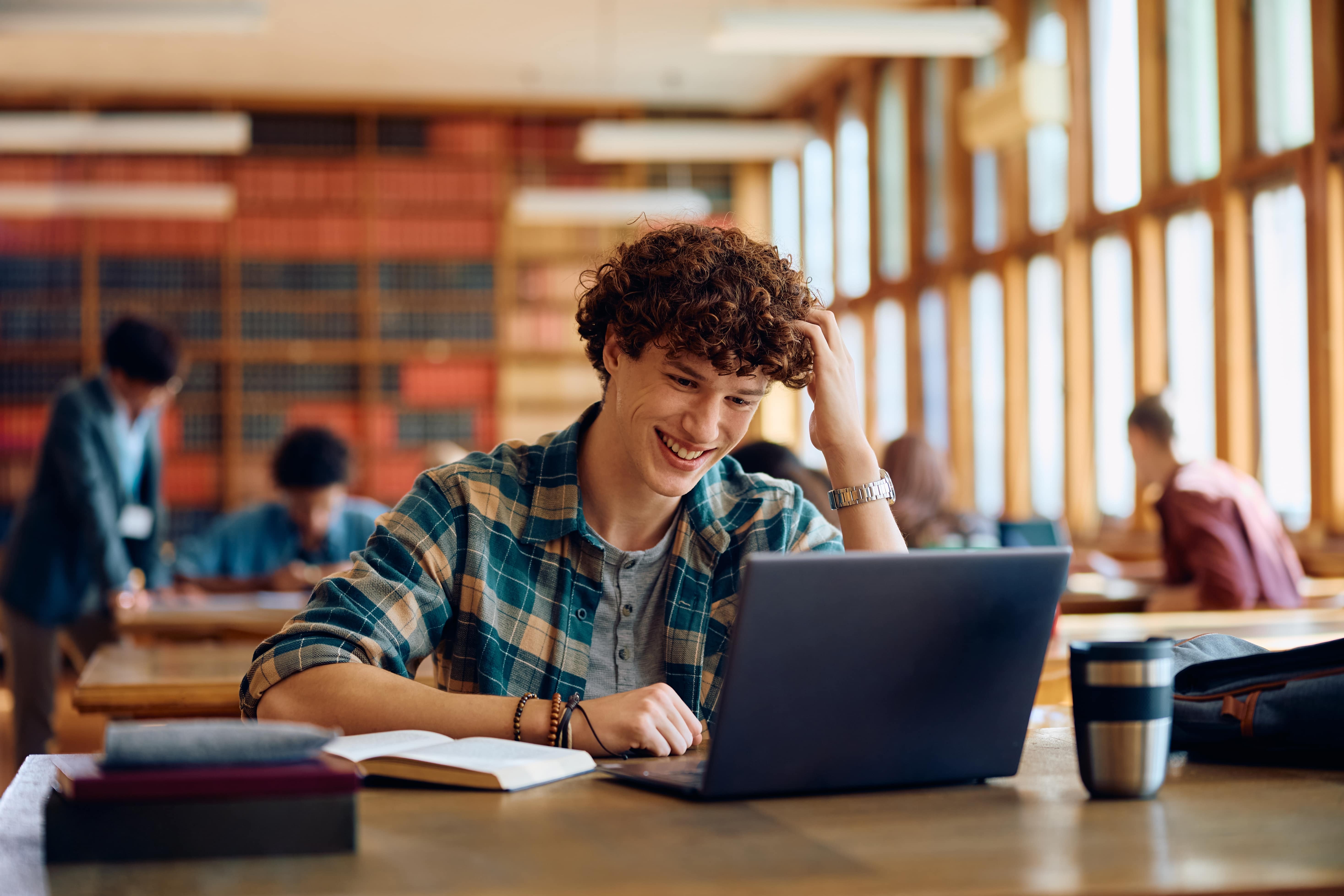 Imagem de um garoto ruivo, aprendendo inglês em seu notebook. Ele está em uma biblioteca e há outros estudantes ao fundo.