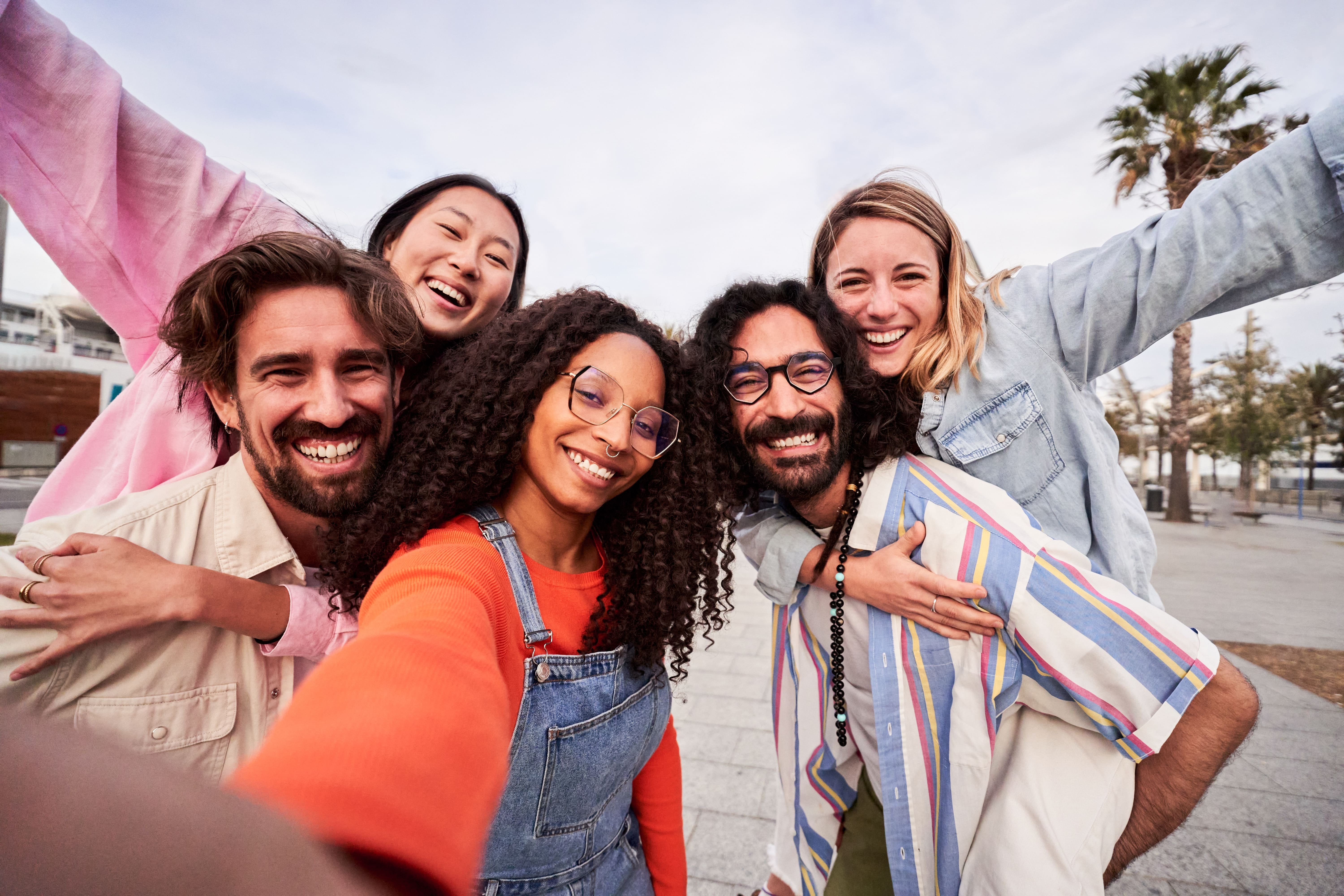 Imagem de um grupo de amigos diverso, com cinco pessoas, sorrindo para uma selfie, em uma praça. 