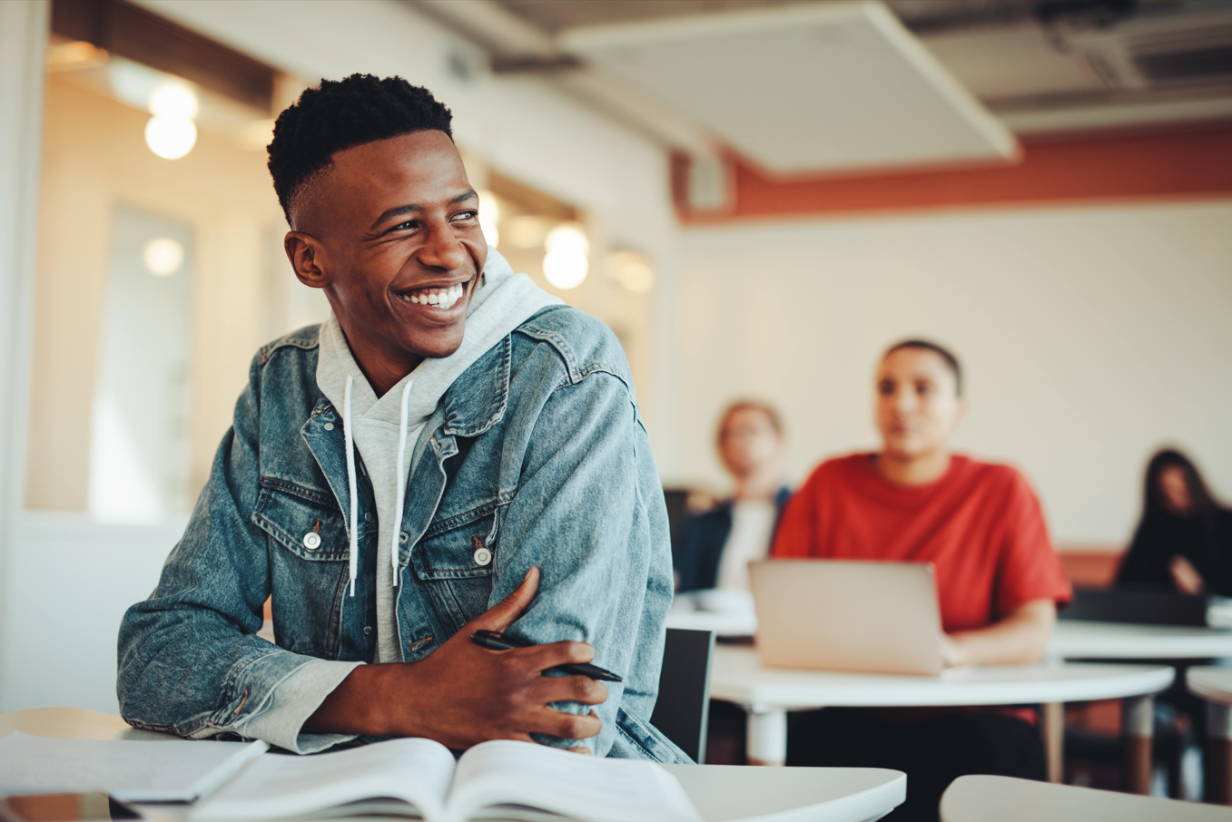 Jovem sorridente em sala de aula, vestindo jaqueta jeans, com livro aberto à sua frente, representando a motivação e o entusiasmo para estudar e alcançar novas oportunidades.