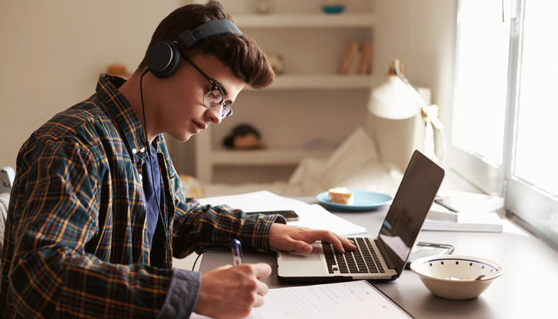 Um jovem estudando em casa com fones de ouvido, concentrado enquanto escreve em um caderno ao lado de seu laptop.