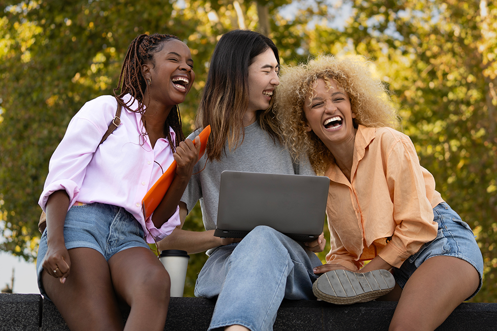 Três jovens estudantes sorrindo, uma delas segura uma pasta e outra um notebook. Estão ao ar livre.