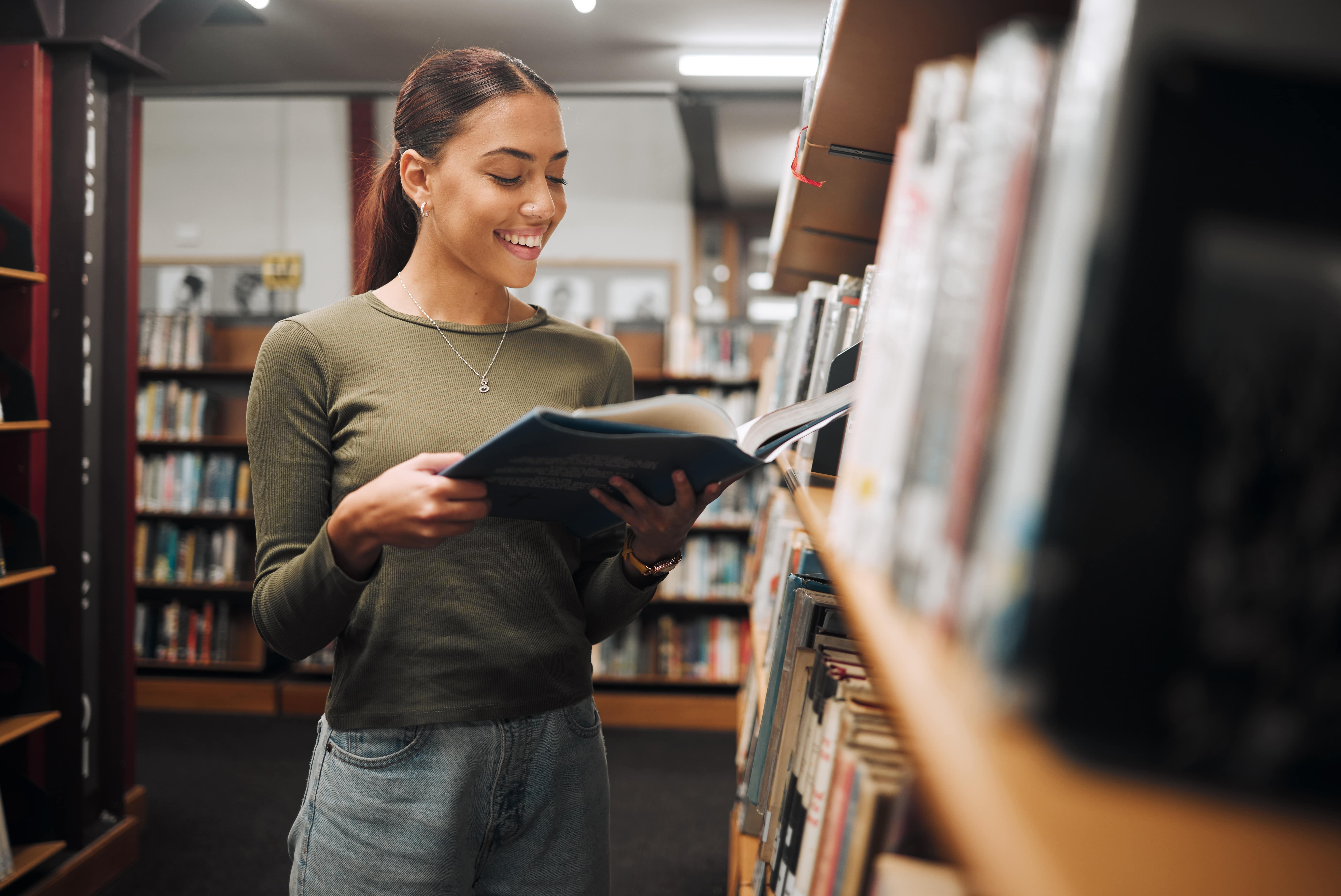 A imagem retrata uma jovem sorrindo enquanto lê um livro em uma biblioteca. Ela está de pé perto de uma estante, vestindo uma blusa verde e jeans. Ela lê para alavancar sua carreira.