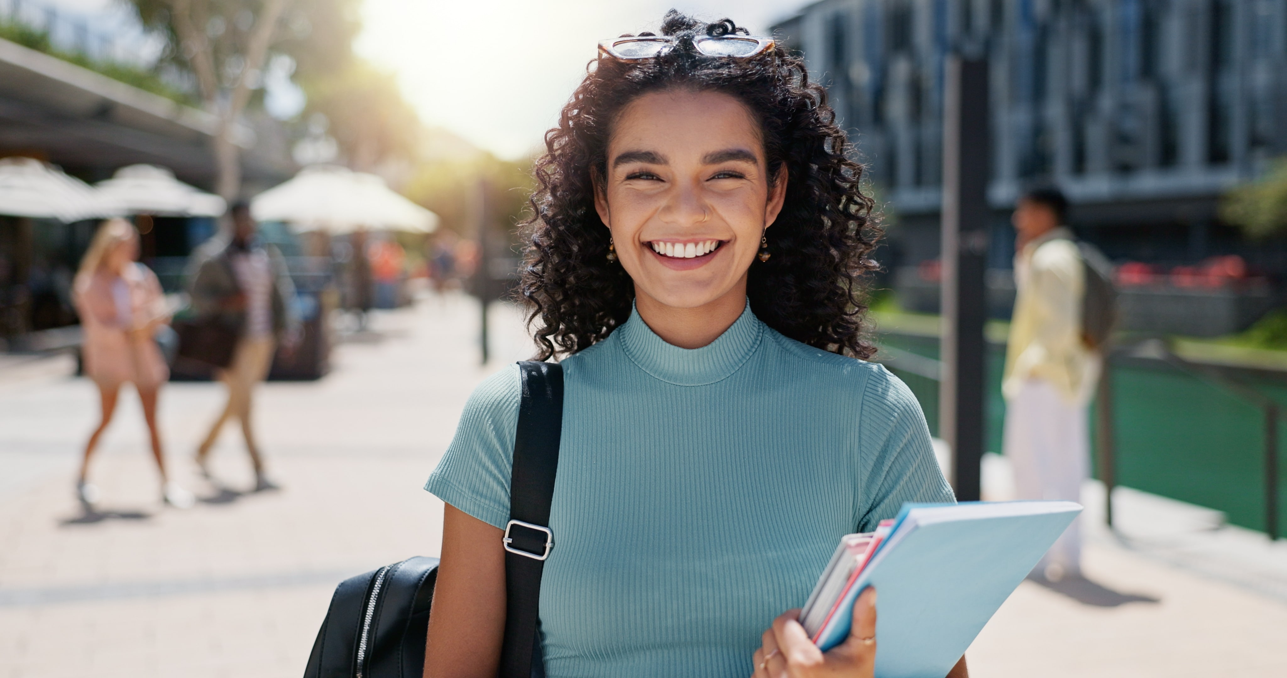 A imagem mostra uma jovem sorridente ao ar livre, carregando cadernos e uma mochila. Ela usa uma blusa verde clara e óculos de sol no cabelo, em um ambiente ensolarado e urbano.