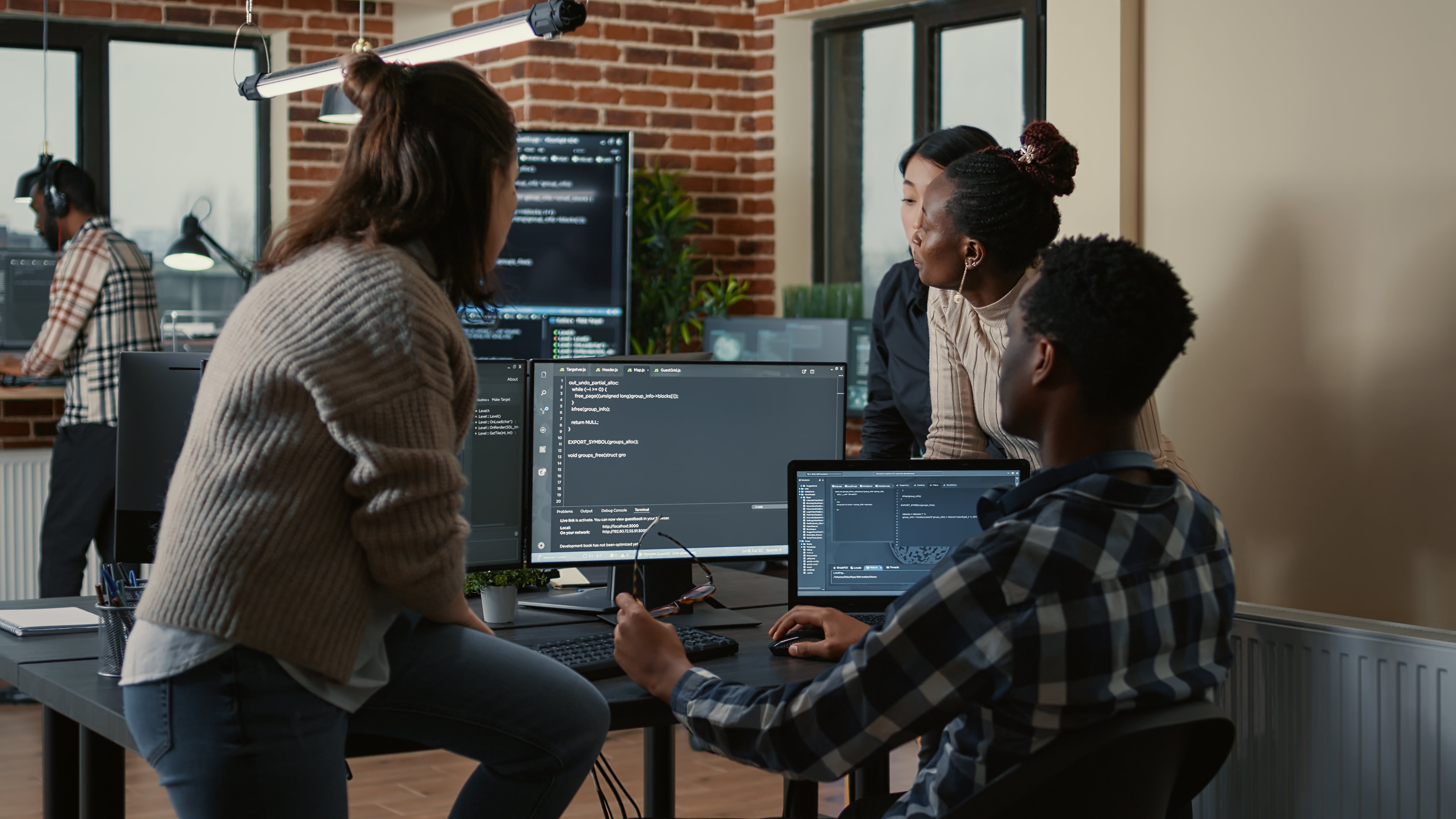 Um grupo de pessoas colabora em um escritório moderno, discutindo códigos exibidos em monitores, destacando trabalho em equipe em um ambiente tecnológico.