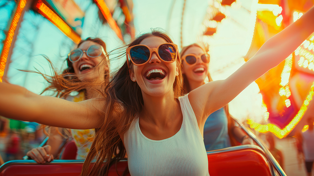 Em primeiro plano uma mulher sorridente com outras duas ao fundo e luzes de uma roda gigante.