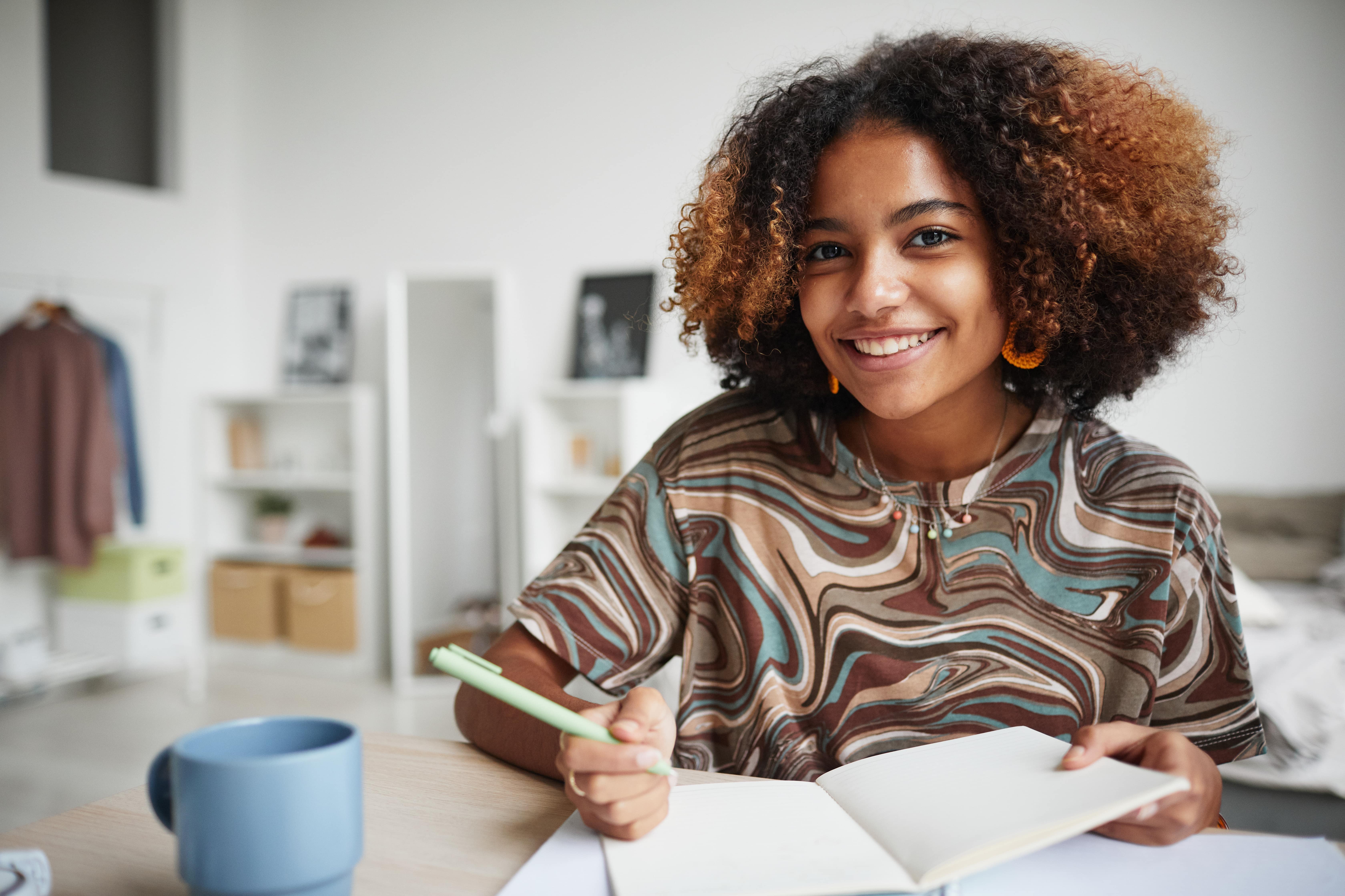 Uma jovem negra sorrindo enquanto segura um caderno e uma caneta. Ao fundo vemos um quarto.