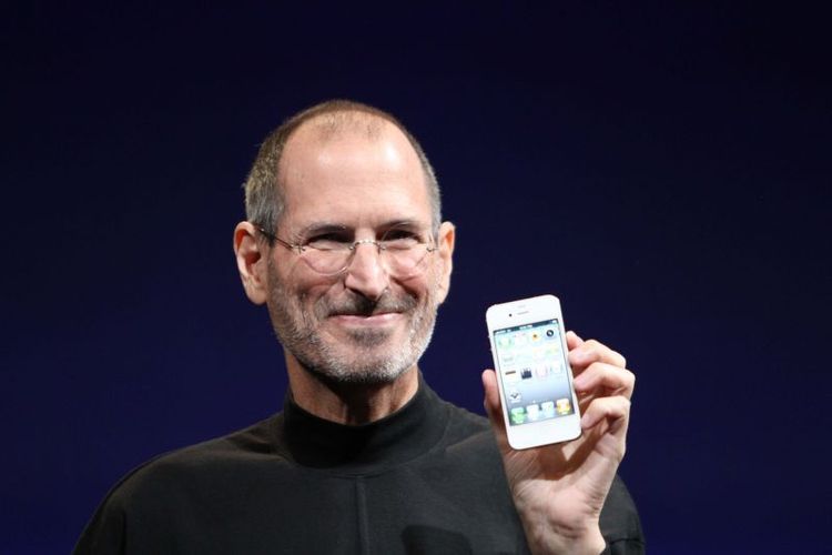 Homem sorrindo enquanto segura um smartphone em destaque, vestindo um suéter preto de gola alta. A imagem simboliza inovação tecnológica e apresentação de um produto icônico.