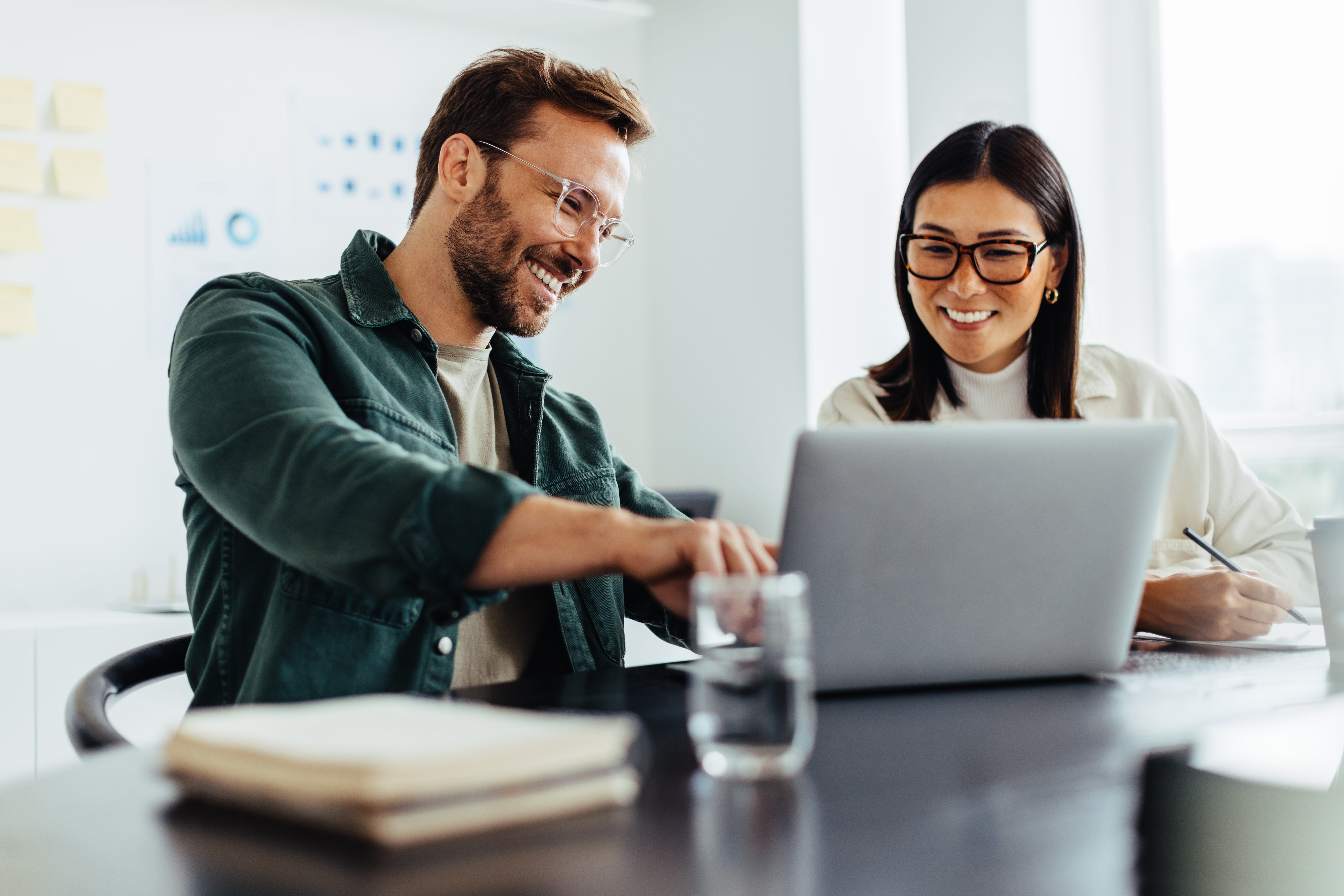 Imagem de um homem e uma mulher, sorrindo em frente a um computador, trabalhando na área de análise e desenvolvimento de sistemas em um escritório. 