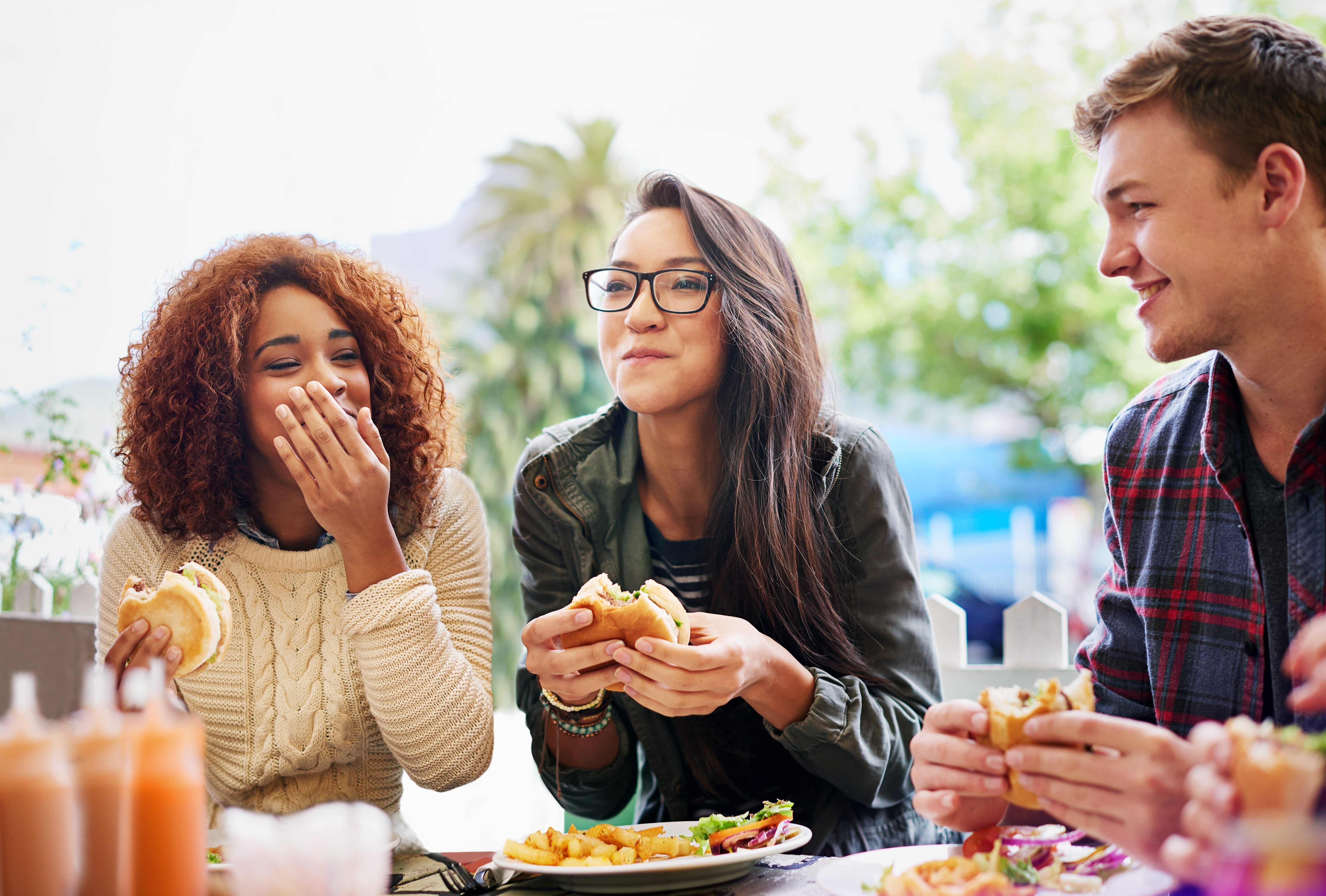 Imagem de um grupo de amigos com três pessoas, comendo hambúrger juntos, eles parecem felizes na situação.