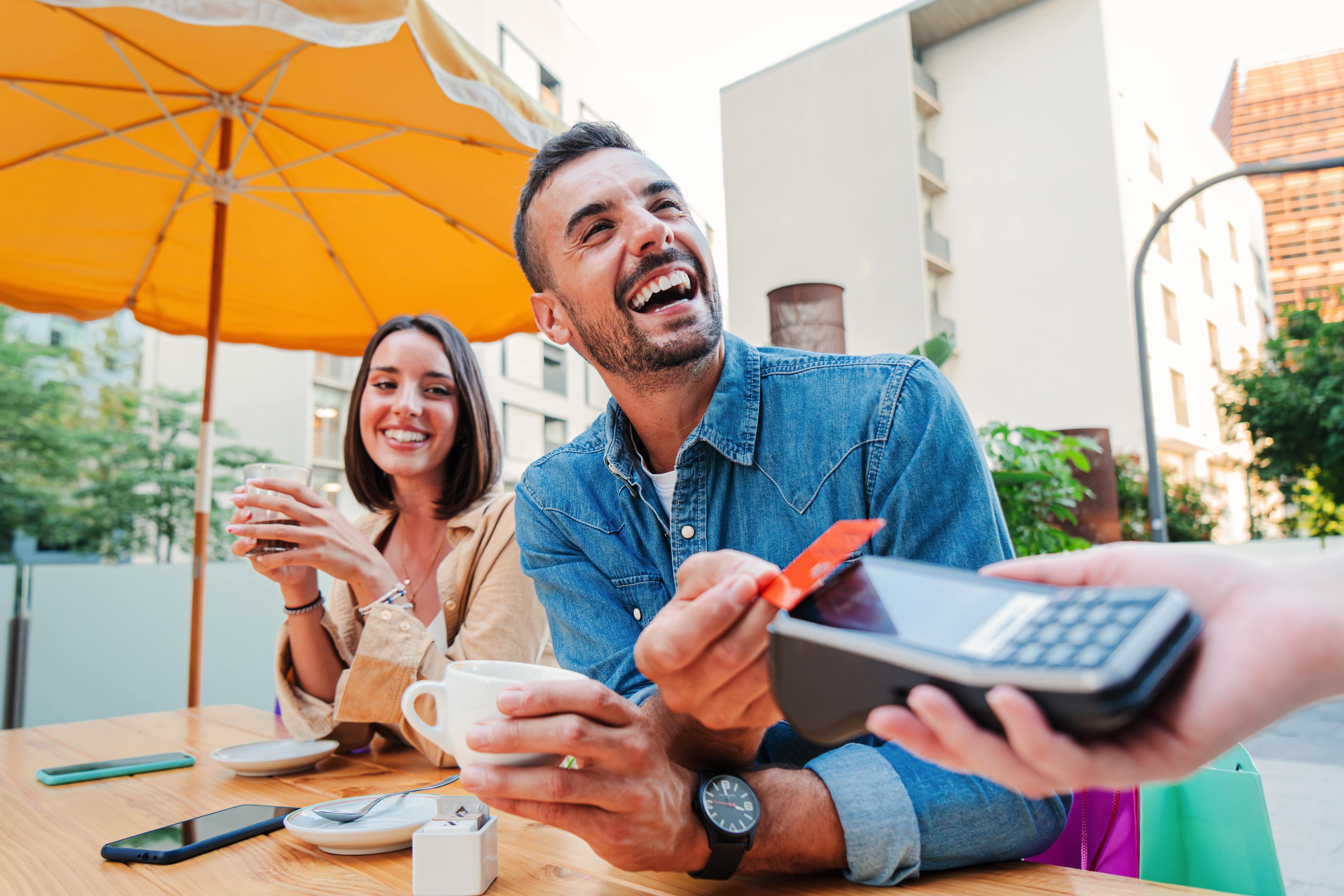Imagem de um casal em um restaurante ao ar livrem eles estão sorrindo enquanto pagam a conta com um cartão. 
