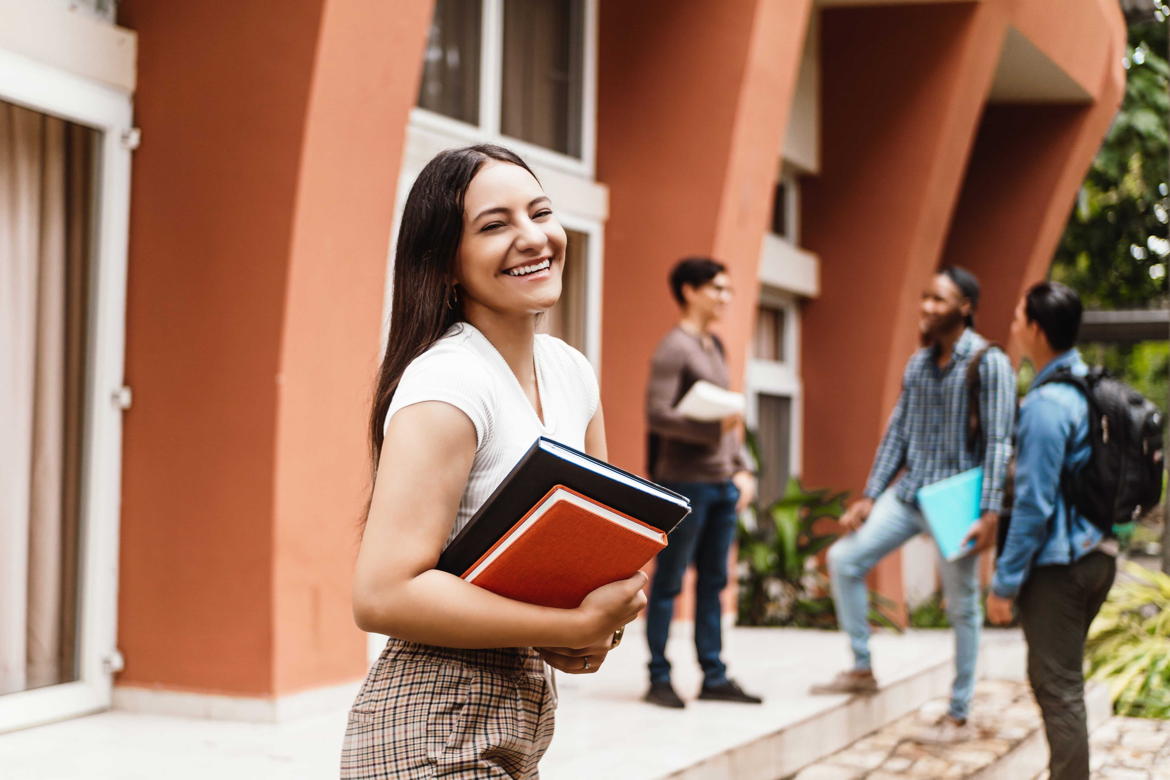 Uma jovem sorri enquanto segura seus cadernos em frente a uma instituição de ensino. Ao fundo, um grupo de colegas conversa. O ambiente inspira motivação e crescimento, refletindo a importância dos cursos profissionalizantes para oportunidades no mercado.