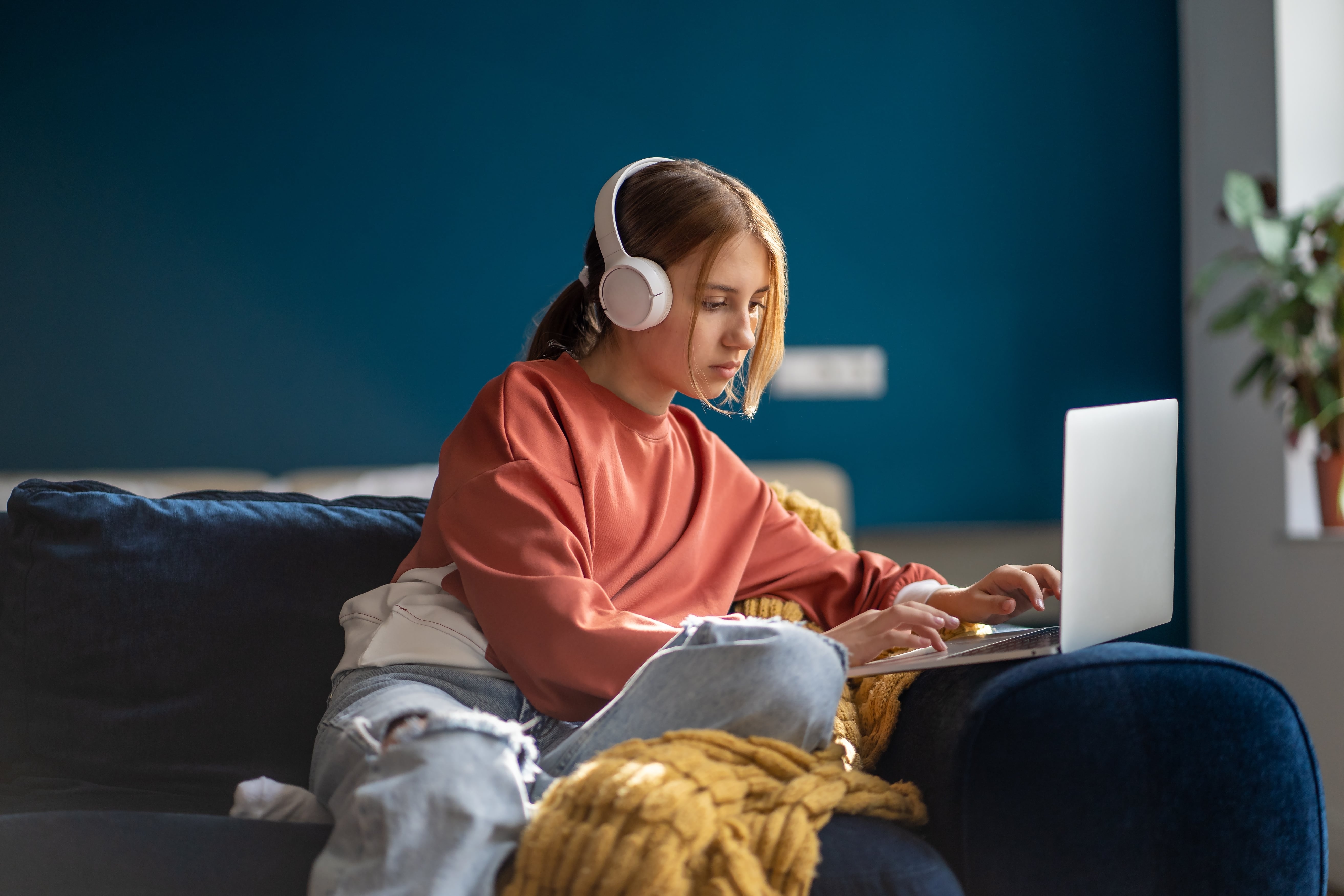 Imagem de uma garota sentada em um sofá azul, ela usa fones de ouvidos enquanto pesquisa a diferença entre bacharelado e licenciatura em seu notebook. 