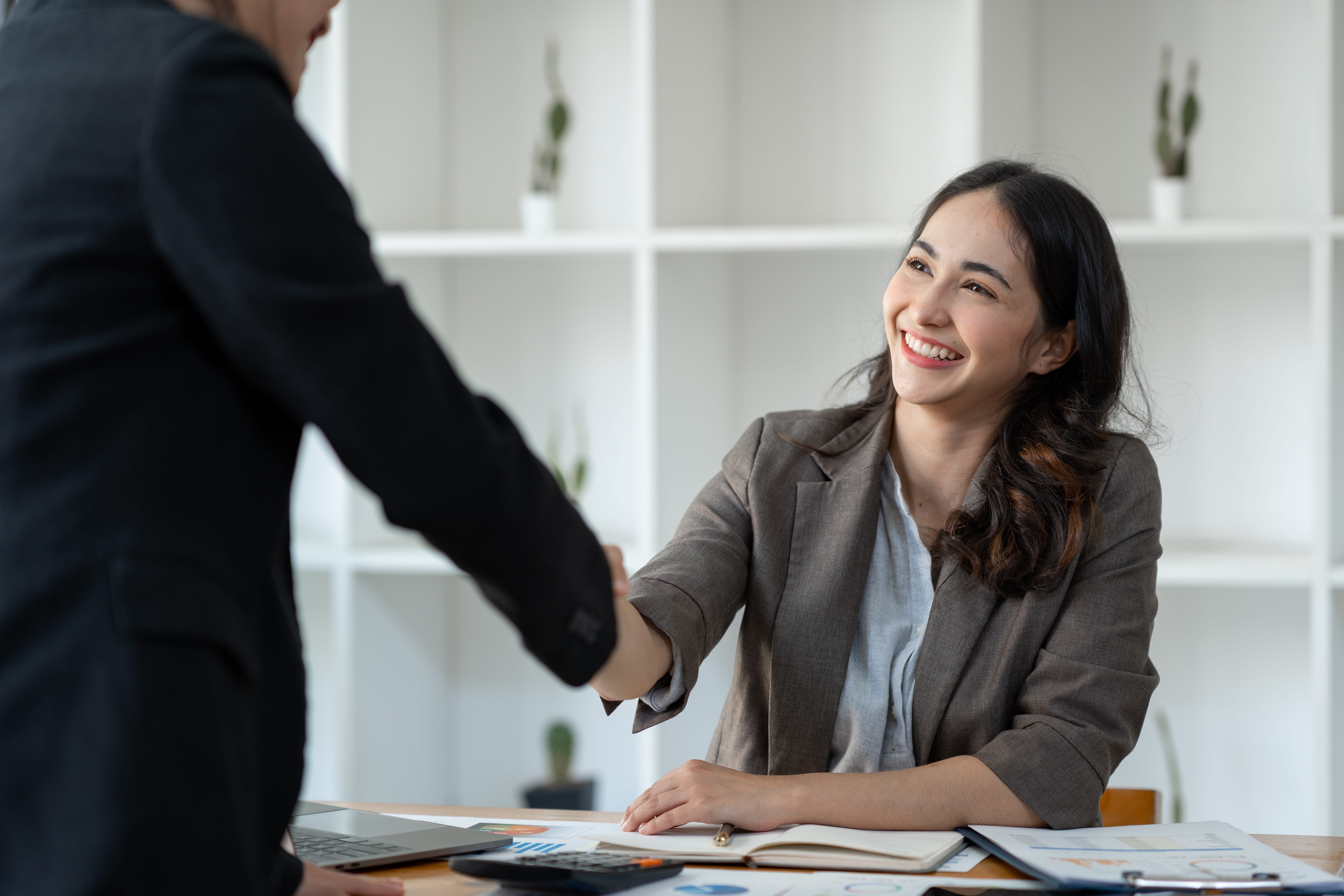 Mulher sorridente, vestida com um blazer cinza, cumprimenta outra pessoa enquanto está sentada em uma mesa de escritório. Ao fundo, prateleiras brancas com pequenos vasos decorativos. A cena transmite uma atmosfera profissional e amigável.