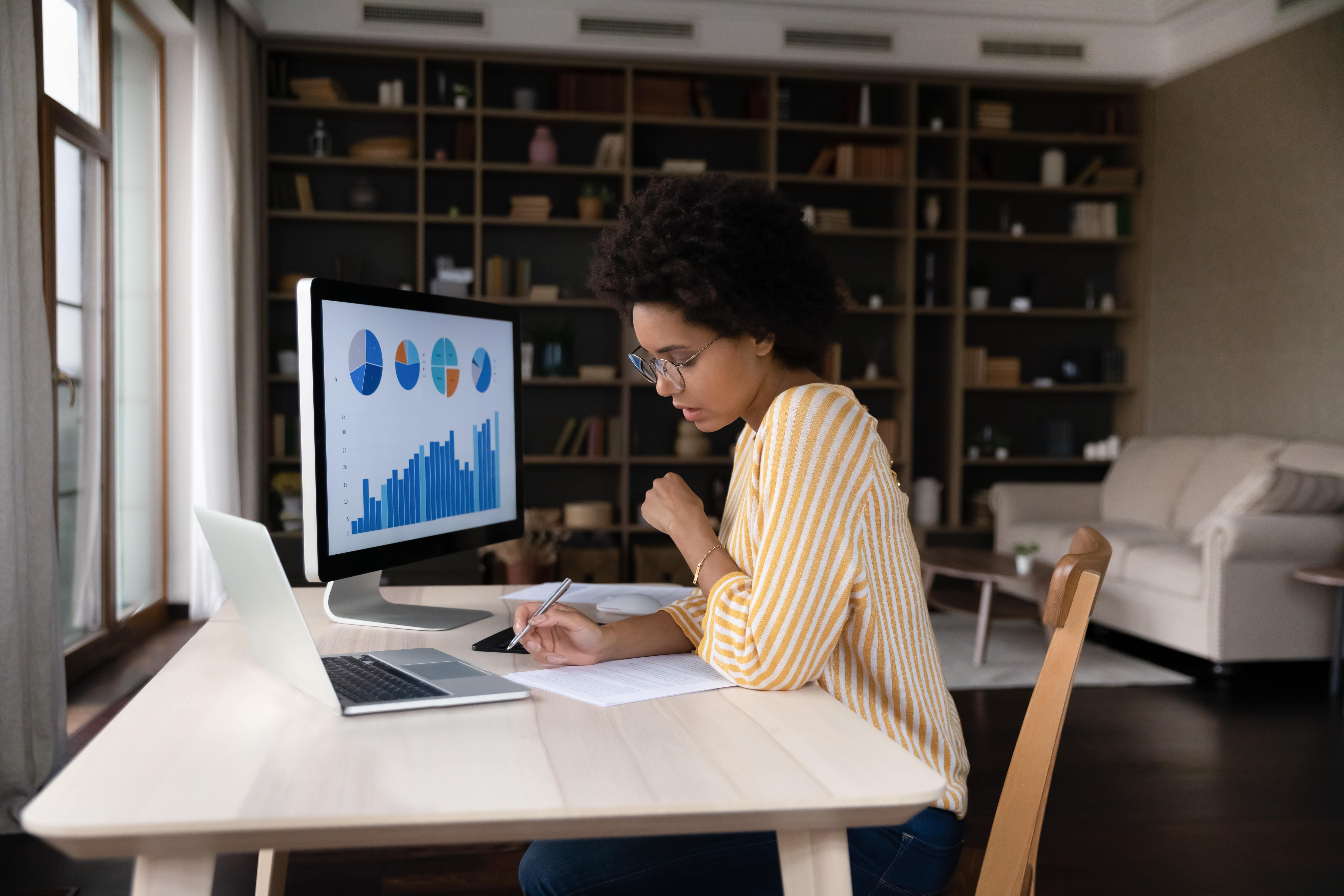 Mulher jovem, de óculos, vestindo uma blusa listrada amarela e branca, trabalha concentrada em um relatório enquanto olha para gráficos e dados exibidos em um monitor de computador. A cena acontece em um ambiente de trabalho home office.