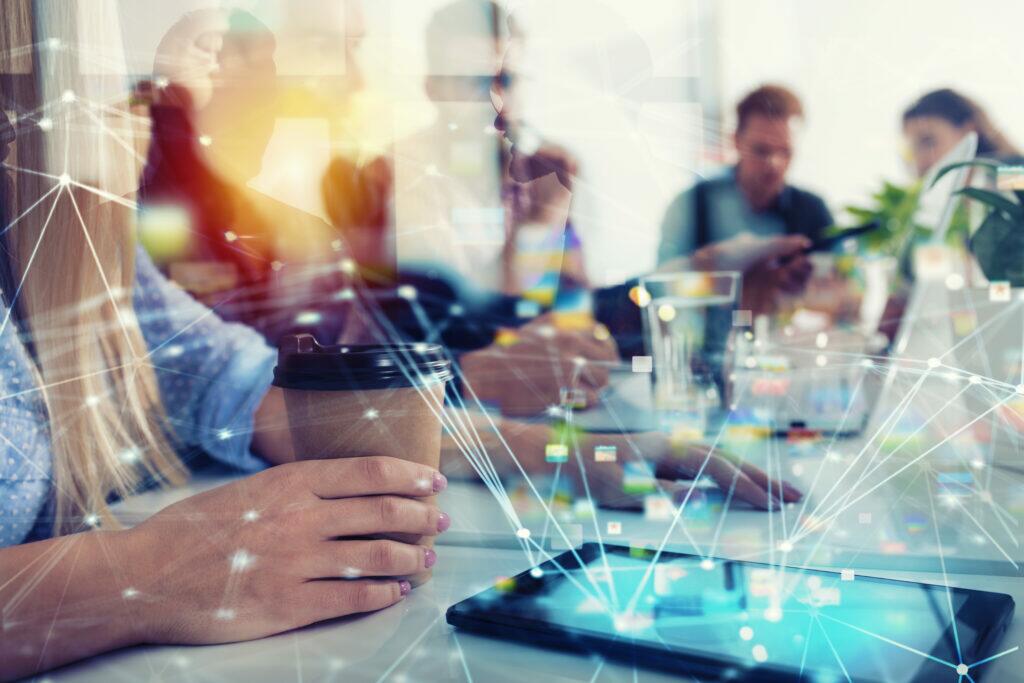 Imagem de pessoas em reunião de negócios, com foco em uma mulher segurando um copo de café, em ambiente de escritório moderno, com discussão e tecnologia presente.