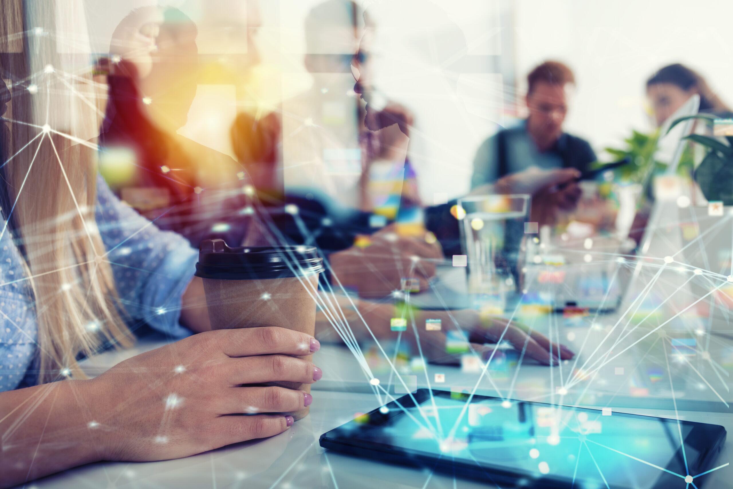 Imagem de pessoas em reunião de negócios, com foco em uma mulher segurando um copo de café, em ambiente de escritório moderno, com discussão e tecnologia presente.