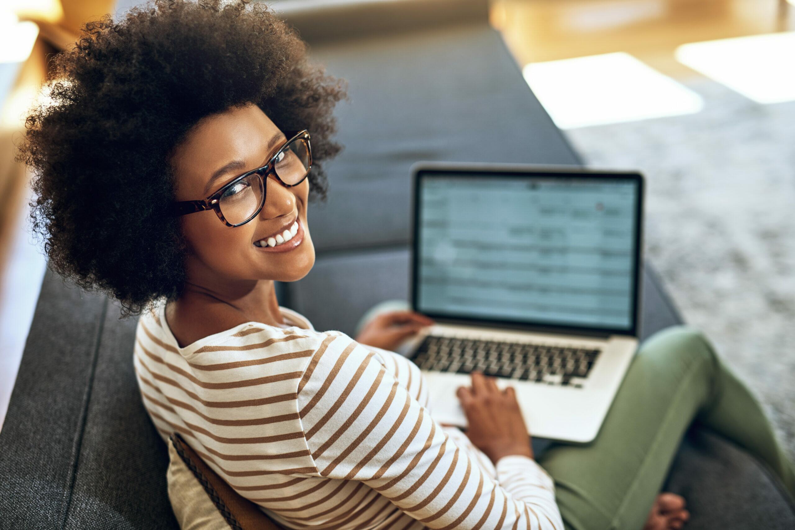 Mulher negra com cabelos encaracolados, usando óculos e camiseta listrada, sorrindo enquanto trabalha em laptop em um ambiente aconchegante.
