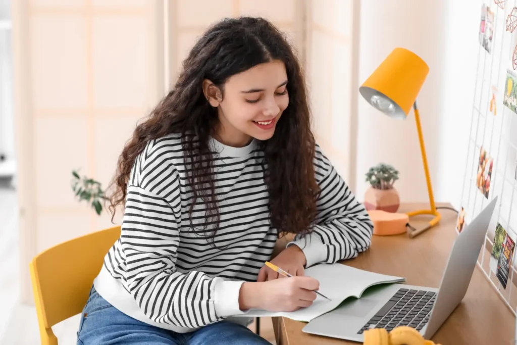 Jovem estudante sorridente estudando em casa com caderno e notebook, incentivando a ideia de nunca parar de estudar para alcançar sucesso e desenvolvimento pessoal.