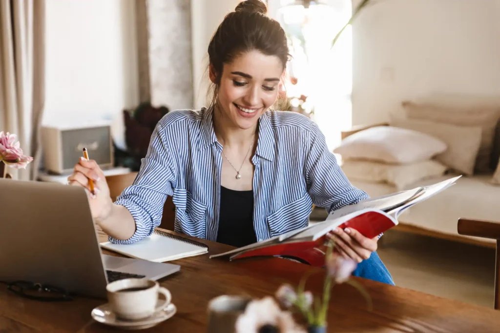 Mulher estudando em casa, com sorriso no rosto, lendo um livro e usando computador portátil para estudar em casa.