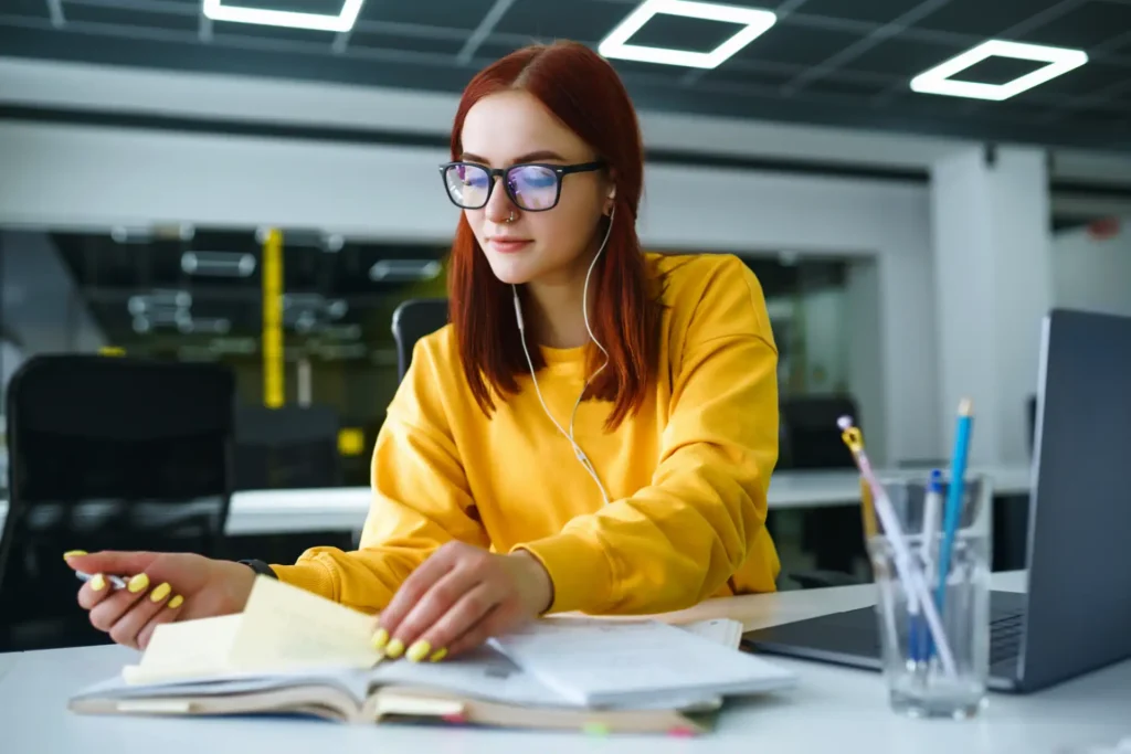 Mulher estudante com cabelo vermelho, usando óculos, na frente de computador, estudando e aproveitando a oportunidade de efetivado no estágio.
