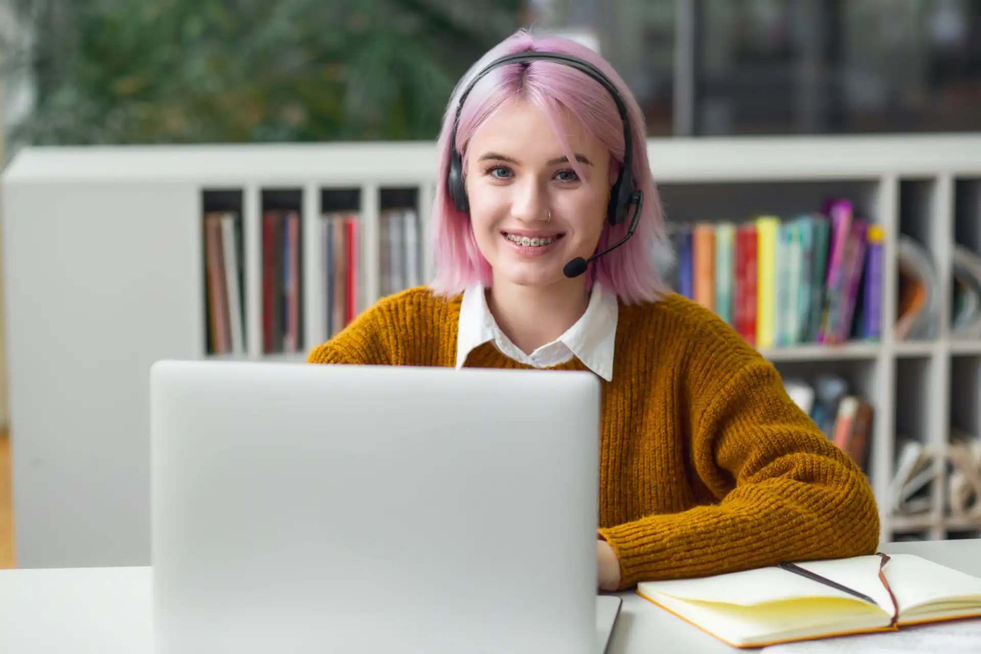 Estagiária de cabelo rosa usando headset, estudando na biblioteca, relacionada à lei do estágio, com sorriso e postura profissional.