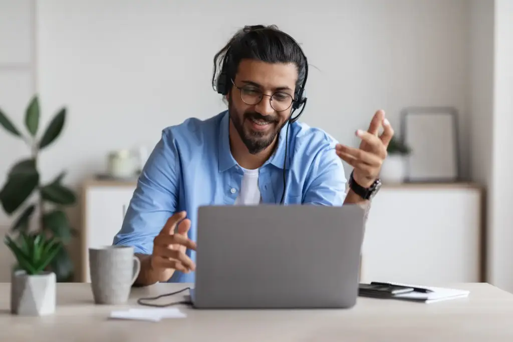 Homem trabalhando em home office usando fones de ouvido, sorrindo e conversando com alguém durante videoconferência, em ambiente de escritório moderno e organizado.
