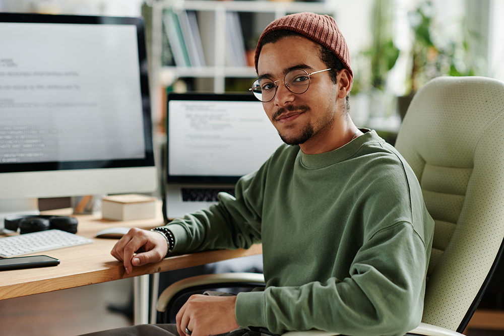 Homem jovem em ambiente de escritório com computadores, usando óculos e suéter verde, sorrindo para a câmera.