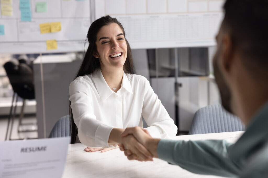 Mulher sorrindo durante entrevista de emprego enquanto aperta a mão de recrutador em escritório moderno, representando dicas sobre o que colocar no objetivo do currículo para primeiro emprego.