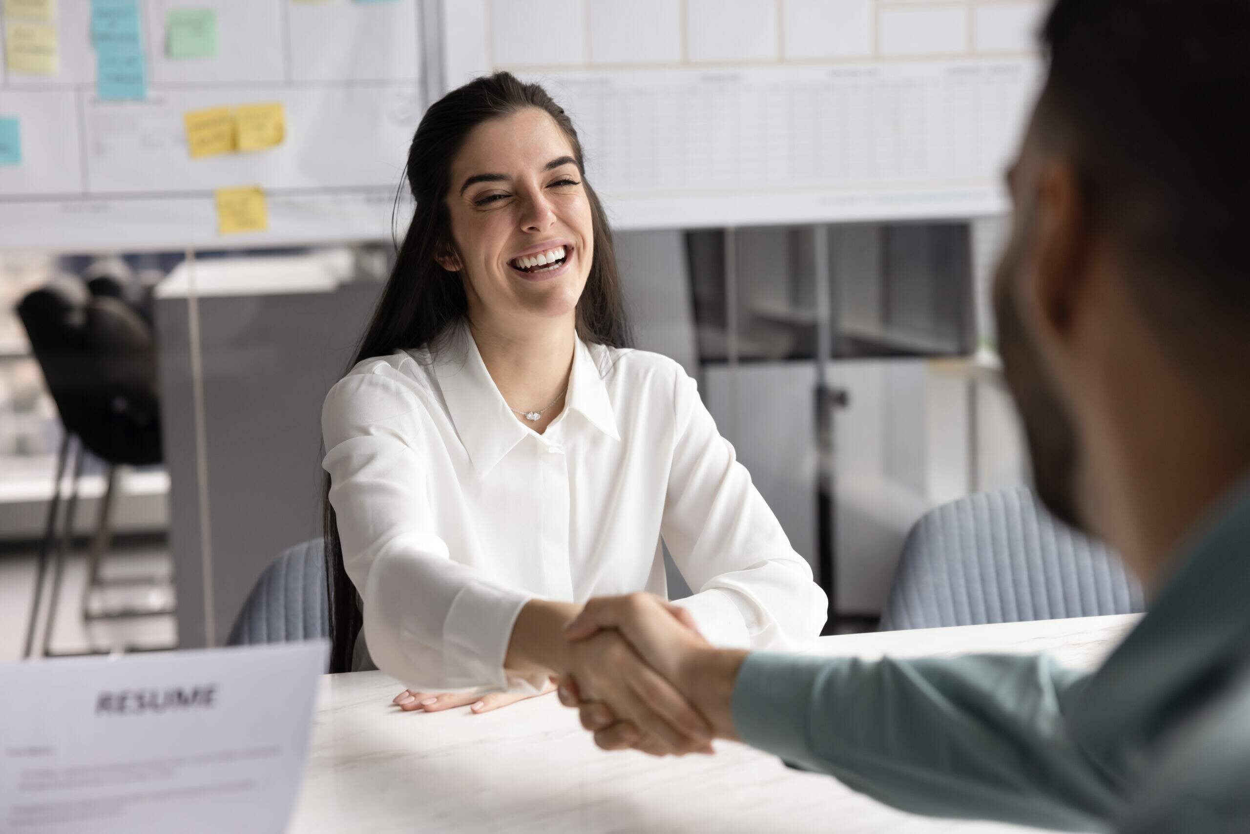 Mulher sorrindo durante entrevista de emprego enquanto aperta a mão de recrutador em escritório moderno, representando dicas sobre o que colocar no objetivo do currículo para primeiro emprego.