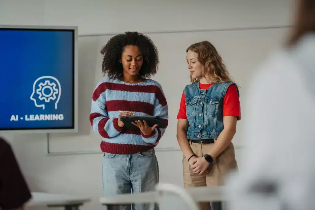 Duas mulheres, uma negra de cabelo crespo e outra loira, em sala de aula participando de um curso de ia, com tela ao fundo mostrando o tema AI LEARNING.