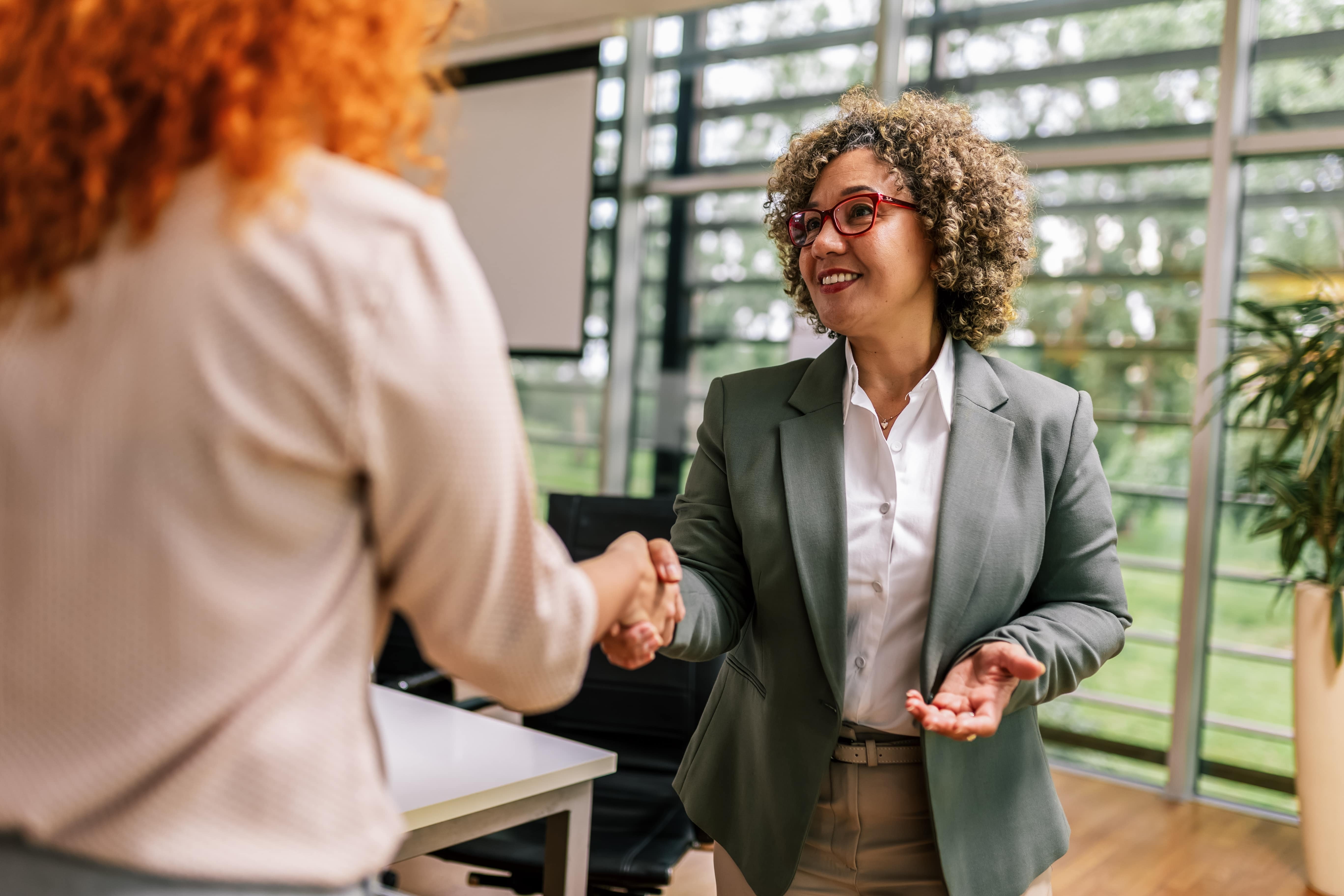 Duas mulheres apertam as mãos em um ambiente de escritório, simbolizando um momento de recrutamento ou negociação dentro do setor de Recursos Humanos. Uma delas está usando óculos vermelhos e um blazer cinza, com um sorriso acolhedor.